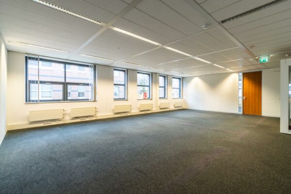 Leegstaand, licht kantoorruimte aan de Lange Dreef met meerdere ramen, radiatoren en een gesloten houten deur.